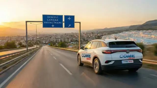 Gün batımında, Denizli Çardak Havalimanı yolunda Pamukkale travertenleri ve şehir manzarası eşliğinde ilerleyen, üzerinde 'COME RENT' logosu bulunan beyaz Volkswagen ID.4 elektrikli araç.
